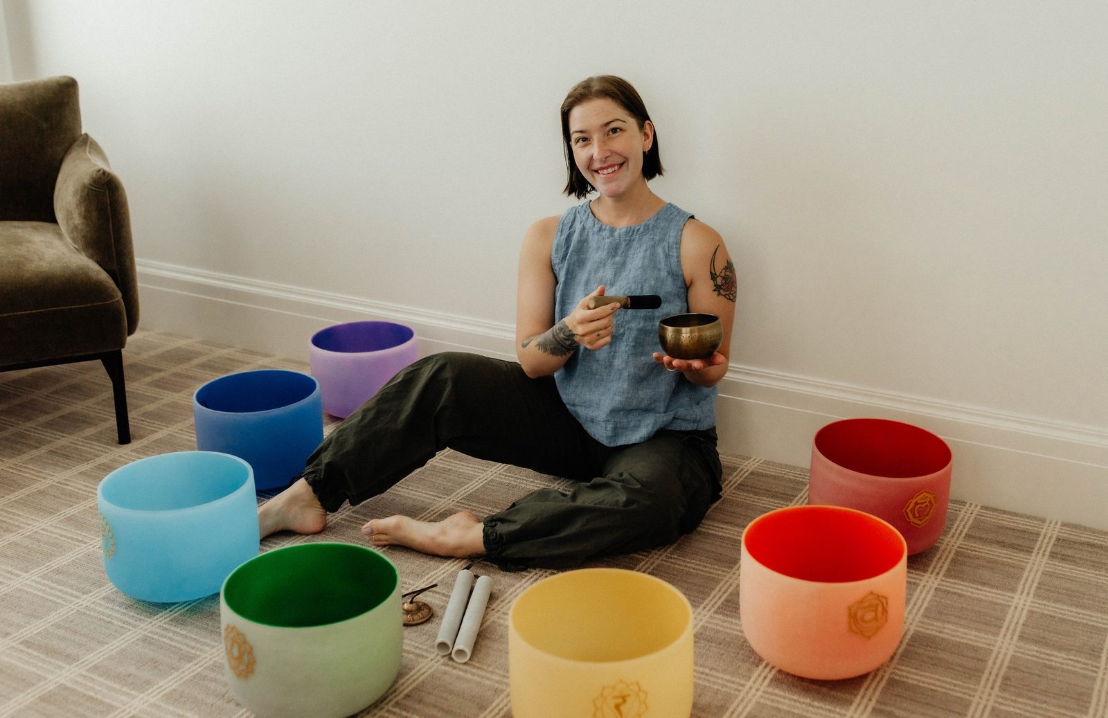 Shelbilee Passey seated on the floor inside a ring of coloured crystal singing bowls, holding a brass bowl and mallet