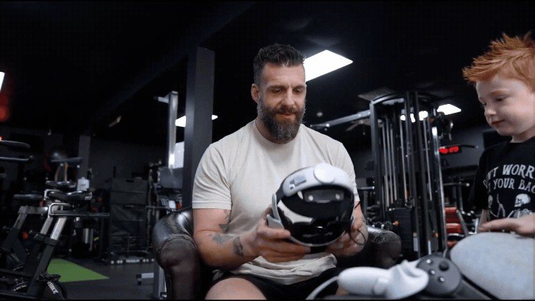 A veteran with prosthetic legs sits in a gym holding a Neurova Labs headset while a young boy watches