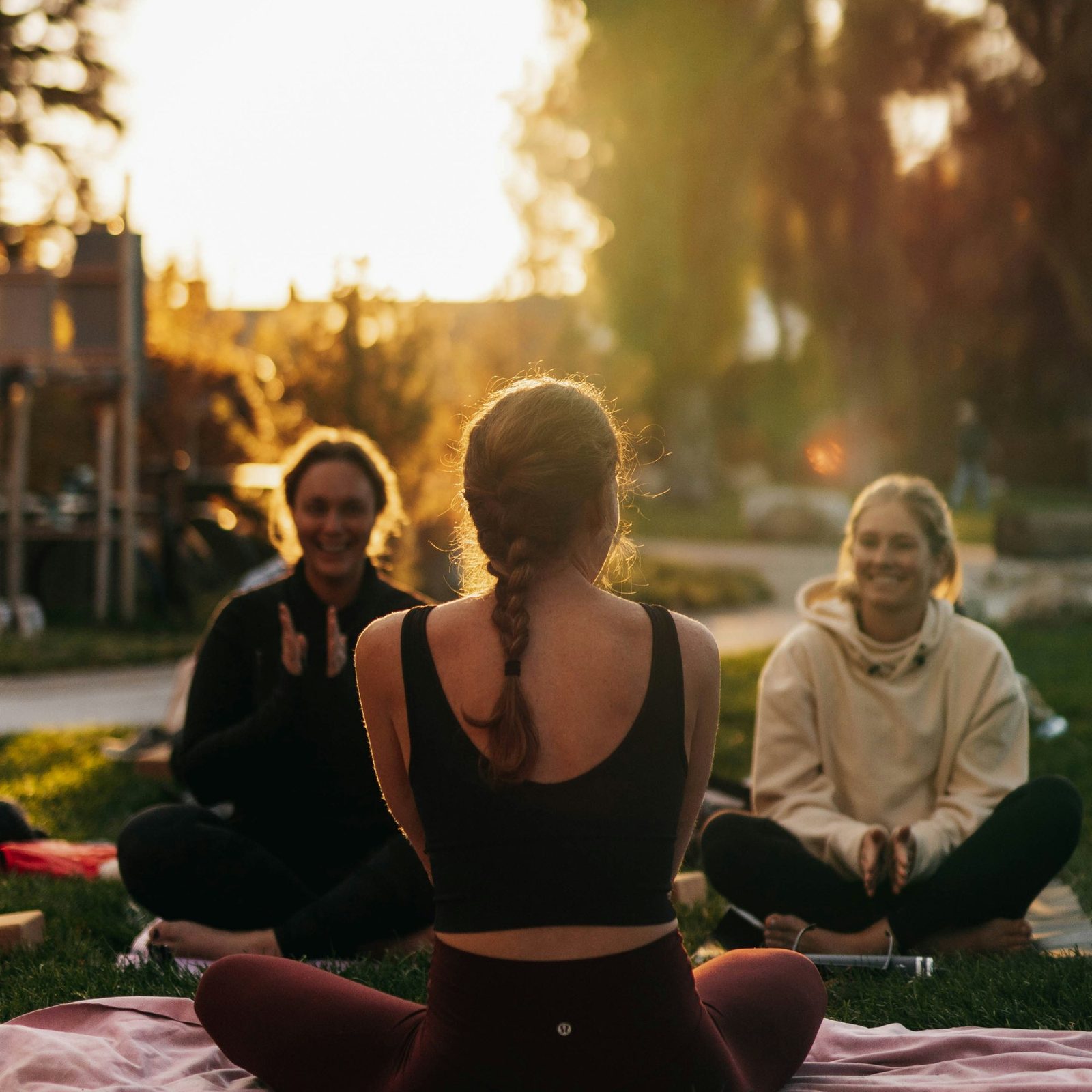 Three women sitting cross-legged on mats outdoors at sunset, smiling together in a yoga session