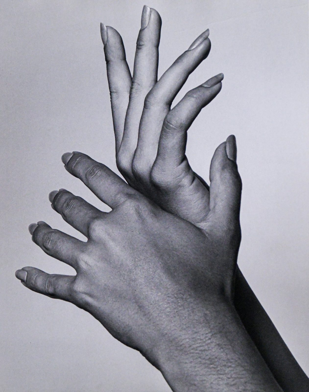 Two hands crossed at the wrists with fingers gracefully outstretched, in a black and white studio photograph against a pale backdrop.