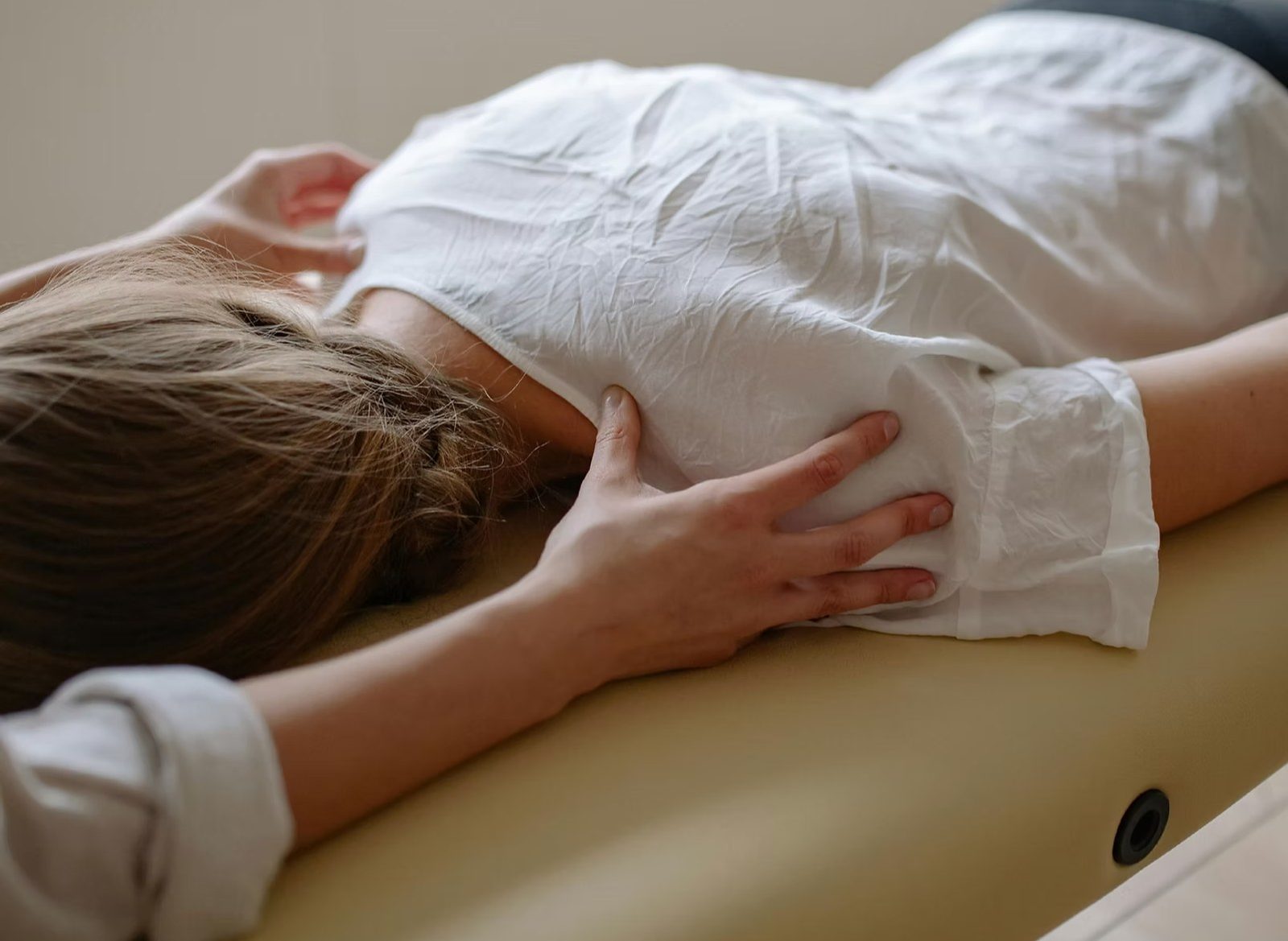 A practitioner's hands resting on the upper back of a woman in a white shirt, lying face down on a treatment table.