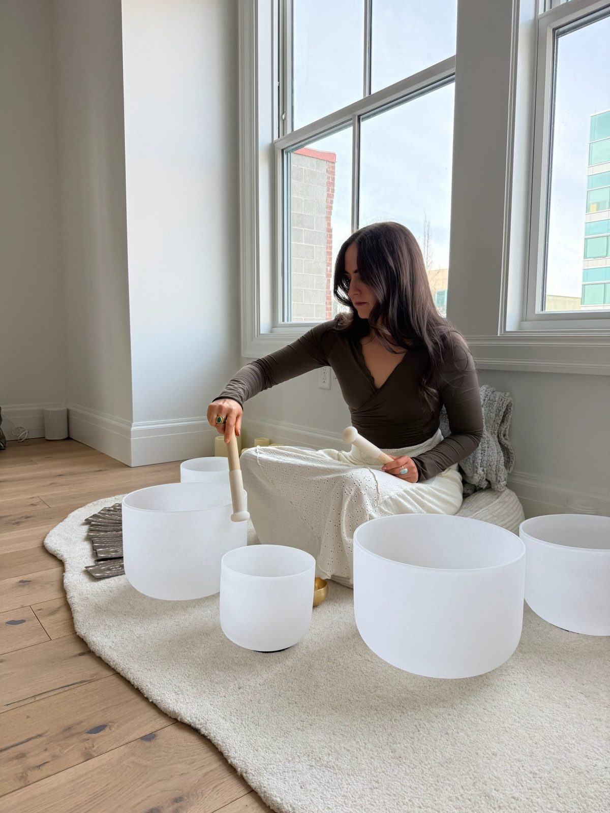 A practitioner sitting on the floor of a sunlit room, playing a circle of crystal singing bowls with a mallet