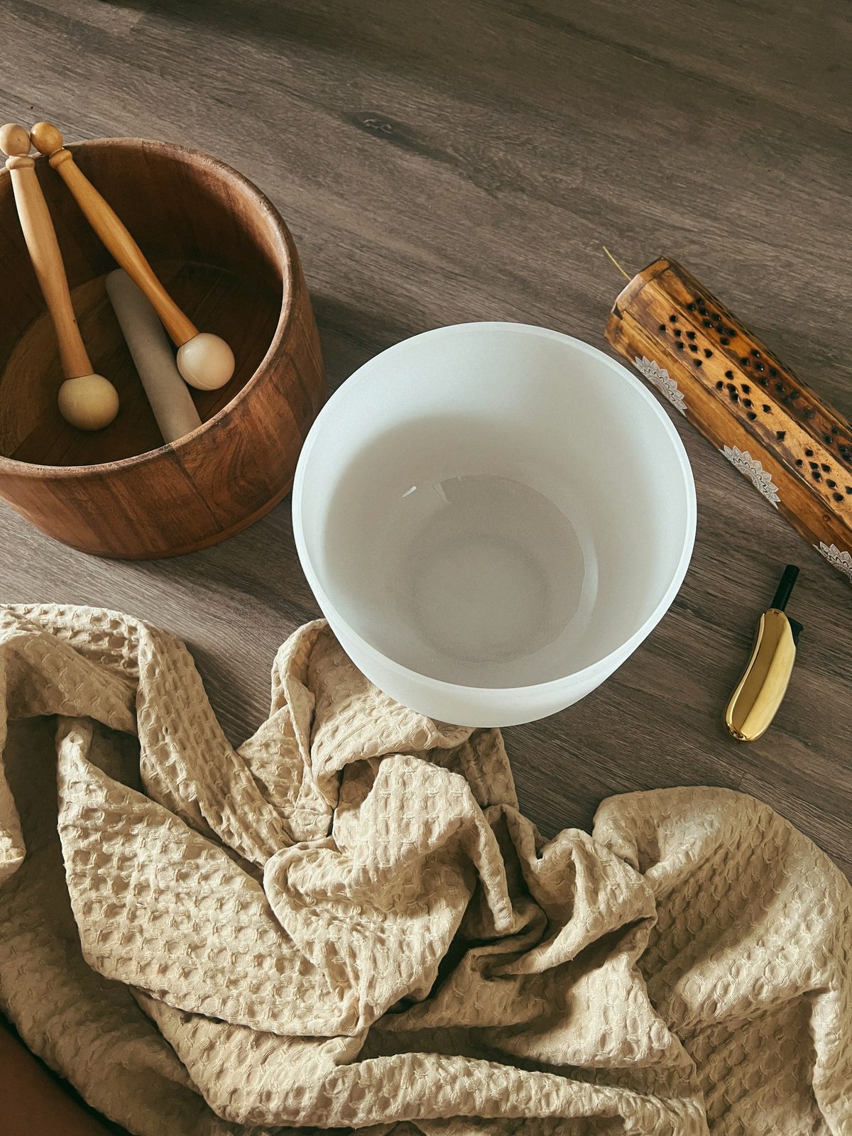 A frosted white crystal singing bowl on a wood floor, beside a wooden bowl of felt-tipped mallets and a rainstick, a carved bamboo incense holder and a folded cream waffle-weave towel.
