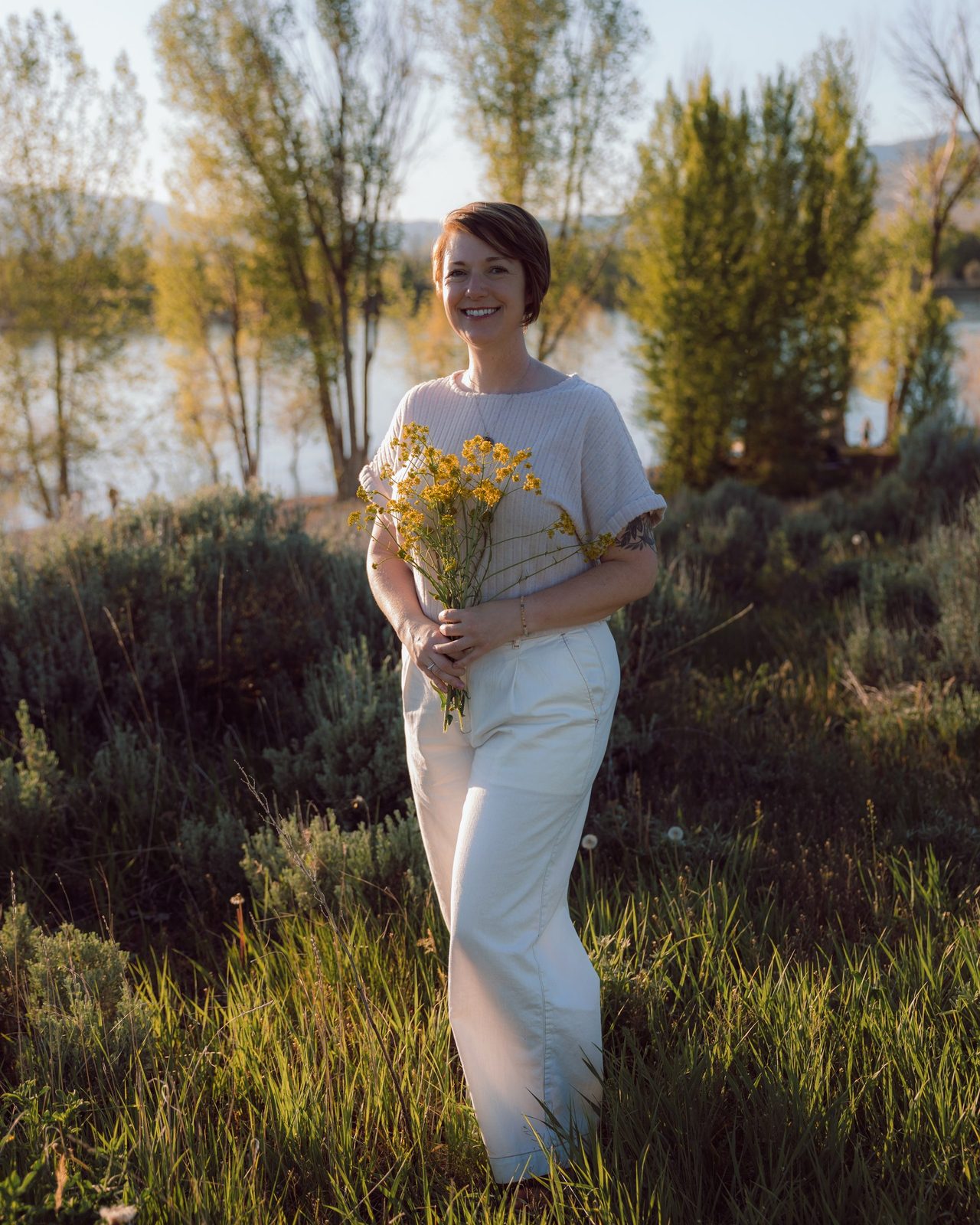 Sarah Tolson holding a bunch of yellow wildflowers in tall grass by a river at golden hour