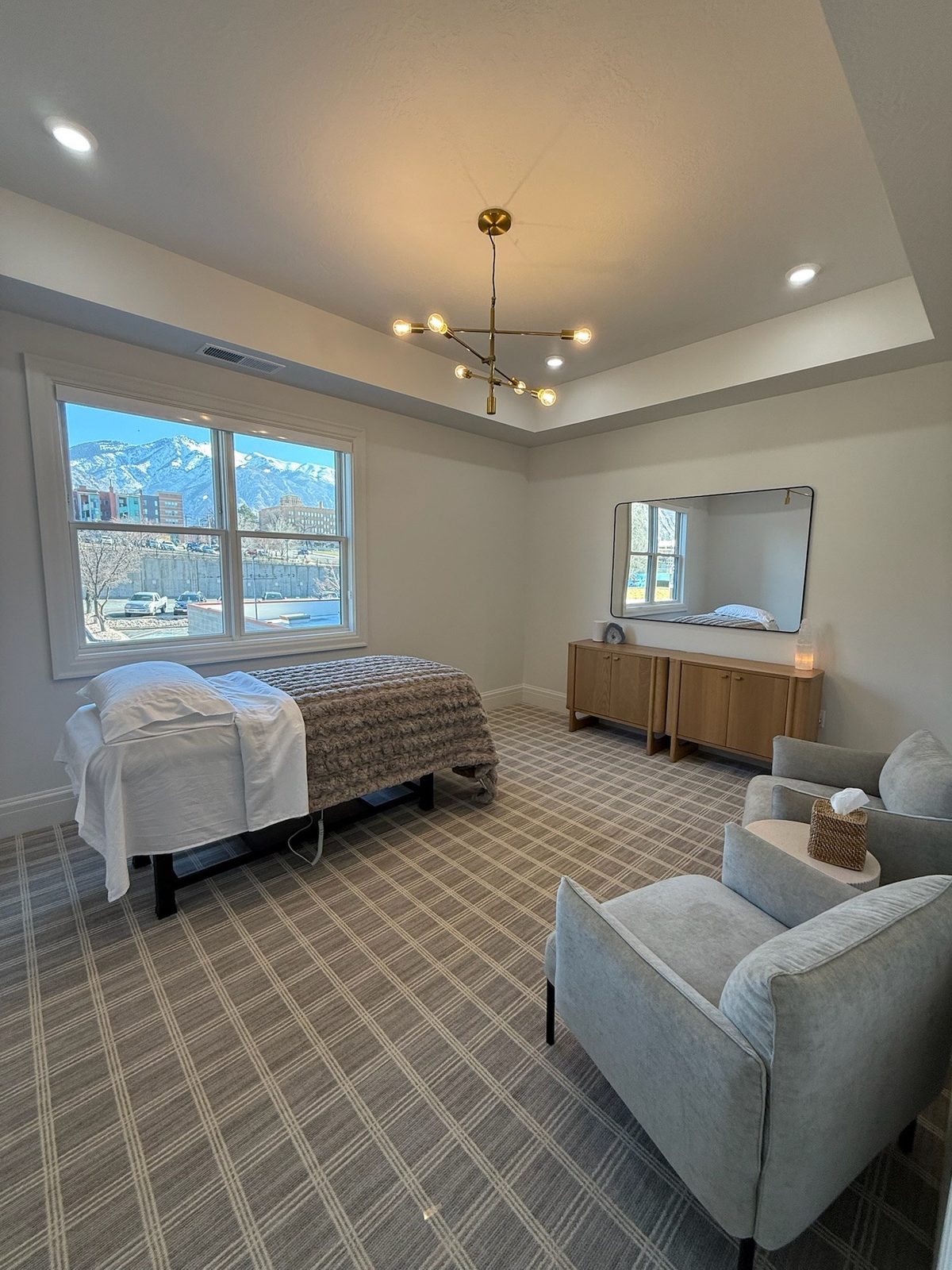 A furnished treatment room: a draped massage table, two soft armchairs, and a snow-topped mountain through the window